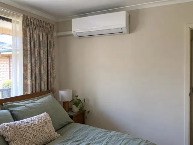 Wall-mounted split system above a bed in a bedroom with curtains and a bedside table in Blakeview